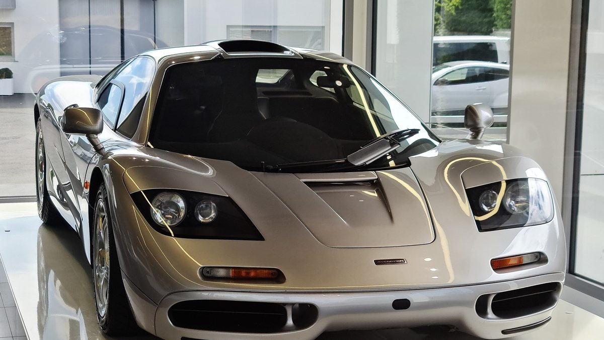A silver McLaren F1 road car with its headlights on, parked on a white showroom floor